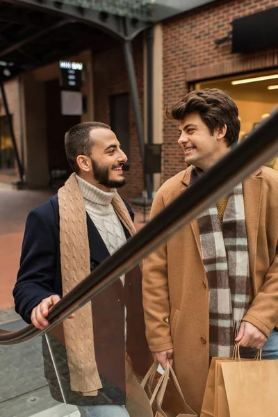 Modischer schwuler Mann im beigen Mantel mit Einkaufstaschen und Blick auf bärtigen Freund auf der Rolltreppe — Stockfoto