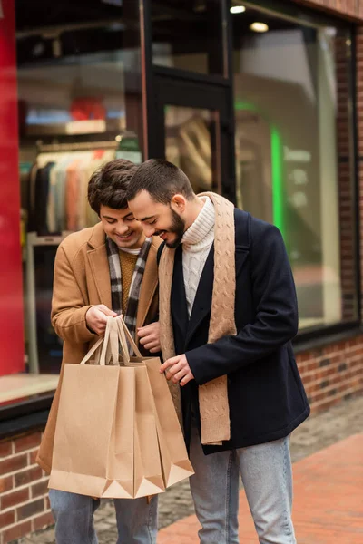 Feliz gay casal no scarfs olhando no compras sacos perto borrado showcase no urbano rua — Fotografia de Stock