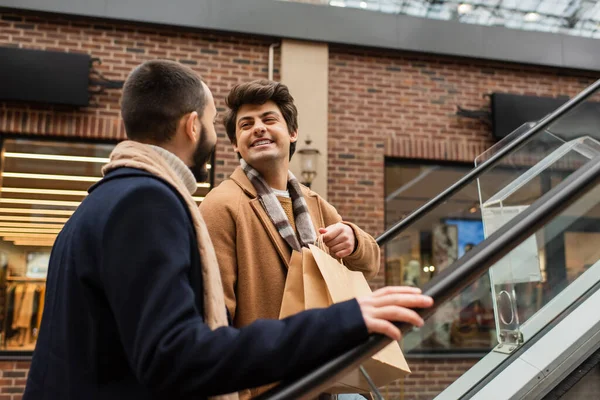 Fröhlicher schwuler Mann mit Einkaufstüten und Blick auf Freund auf Rolltreppe — Stockfoto