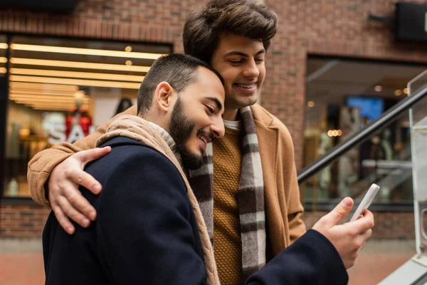Молодий і щасливий гей чоловік в модному вбранні обіймає хлопця зі смартфоном на міській вулиці — стокове фото