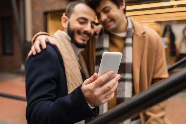 Sorridente gay uomo abbracciando barbuto fidanzato utilizzando cellulare all'aperto su sfondo sfocato — Foto stock