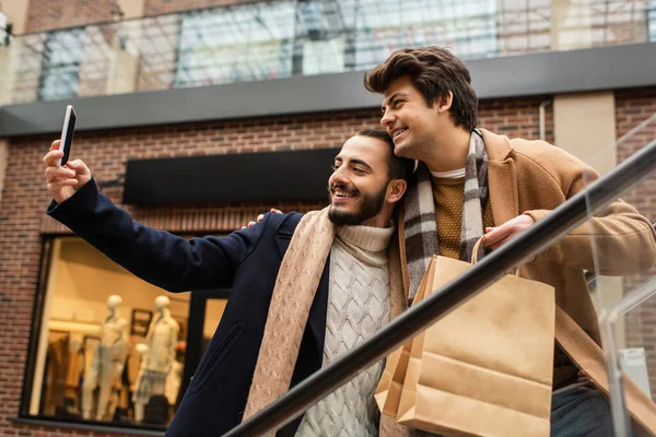 Alegre pareja gay en moda abrigos y bufandas tomando selfie en smartphone en escaleras mecánicas - foto de stock