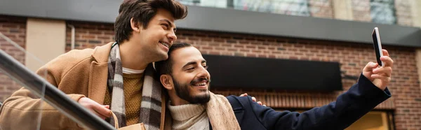Bearded gay man taking selfie with boyfriend holding shopping bag outdoors, banner — Stock Photo