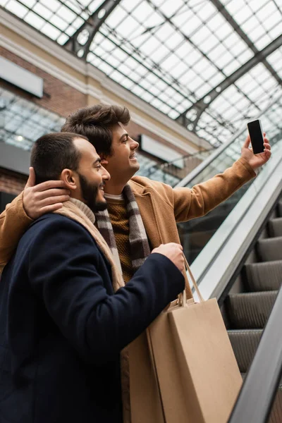 Young man hugging neck of bearded and trendy boyfriend with shopping bags and taking selfie on escalator — Stock Photo