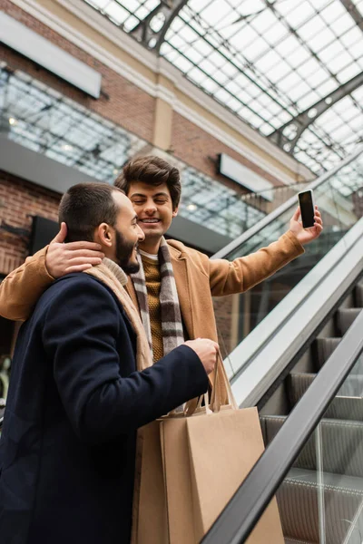 Joven y feliz hombre abrazando cuello de novio con bolsas de compras mientras toma selfie en escaleras mecánicas - foto de stock