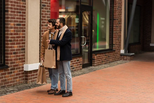 Comprimento total de barbudo gay homem com compras sacos apontando para showcase perto jovem namorado — Fotografia de Stock