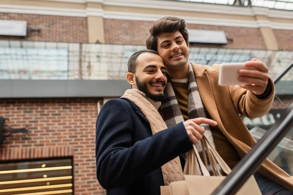Hombre barbudo sonriente con bolsas de compras apuntando al teléfono móvil en la mano del novio - foto de stock