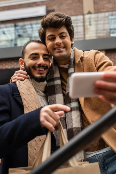 Улыбающийся гей обнимает бородатого парня с сумками для покупок, делая селфи на размытом мобильном телефоне — стоковое фото