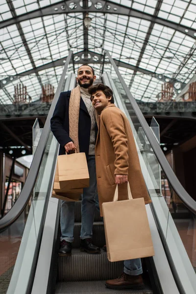 Low angle view of cheerful gay men in stylish casual clothes holding shopping bags and looking away on escalator — Stock Photo