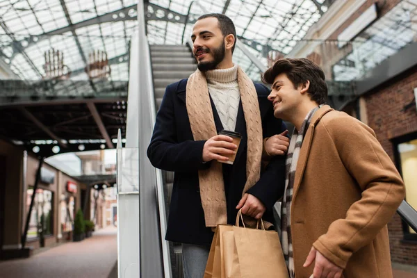 Hombre barbudo con bolsas de compras y taza de papel mirando hacia otro lado cerca novio sonriente en escaleras mecánicas - foto de stock