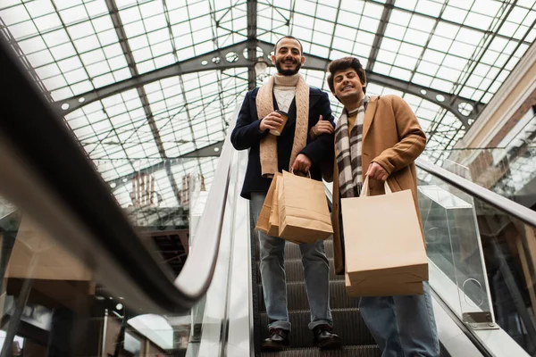 Low angle view of stylish gay partners with coffee to go and shopping bags smiling on escalator — Stock Photo