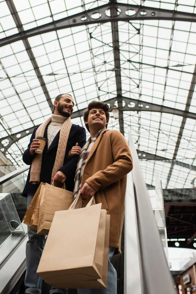 Vista de ángulo bajo de la pareja gay de moda y feliz con bolsas de compras y taza de papel mirando hacia otro lado en escaleras mecánicas - foto de stock