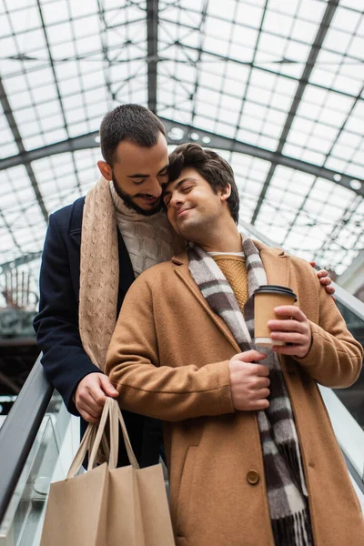 Щасливий гей чоловік з кавою, щоб піти стояти з закритими очима біля бородатого хлопця з сумками на ескалаторі — стокове фото