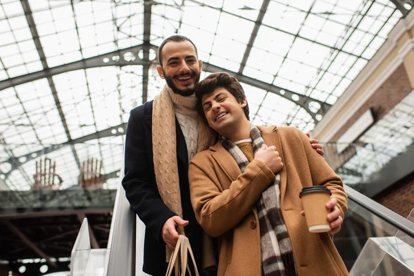 Basso angolo vista di felice gay uomo con caffè per andare vicino ridere barbuto fidanzato su scala mobile — Foto stock