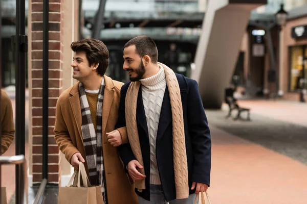 Hombres gay en trajes de moda sosteniendo bolsas de compras mientras mira escaparate en la calle urbana - foto de stock