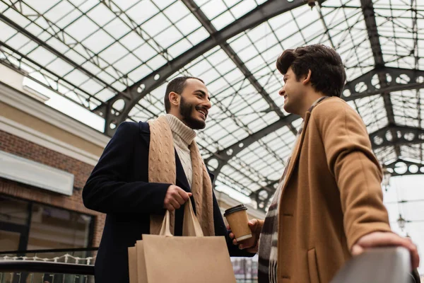 Low angle view of stylish homosexual couple with shopping bags and paper cup under transparent roof outdoors — Stock Photo