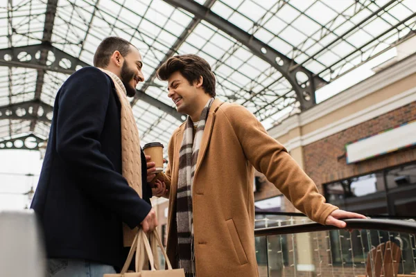 Cheerful gay holding paper cup and laughing near bearded boyfriend with shopping bags under transparent roof — Stock Photo