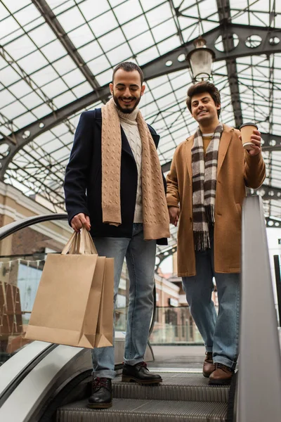 Sonriendo gay los hombres en de moda ropa de pie con compras bolsas y papel taza en escaleras mecánicas - foto de stock