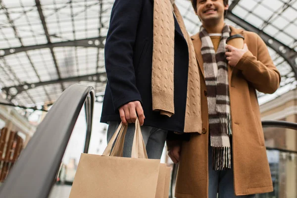 Cropped view of trendy gay man with shopping bags near blurred boyfriend with takeaway drink on escalator — Stock Photo