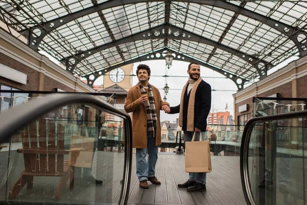 Full length of trendy gay couple with shopping bags and coffee to go holding hands under transparent roof near escalator — Stock Photo