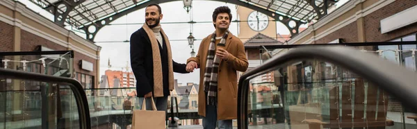 Joyful and stylish gay men with shopping bags and coffee to go holding hands on city street, banner — Stock Photo