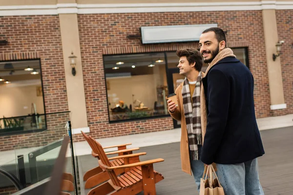 Stylish gay men with shopping bags and coffee to go standing near benches and shops on urban street — Stock Photo
