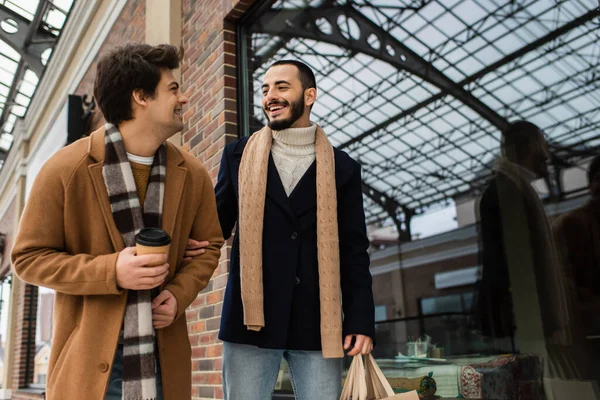 Cheerful and stylish gay couple with paper cup and shopping bags smiling near showcase — Stock Photo