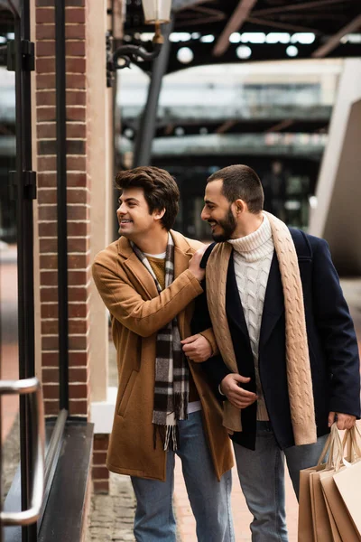Cheerful and trendy gay men with shopping bags looking at showcase on city street — Stock Photo