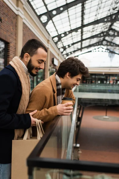 Vista lateral de casal gay sorridente com sacos de compras e café para ir olhando para baixo perto de cerca de vidro ao ar livre — Fotografia de Stock
