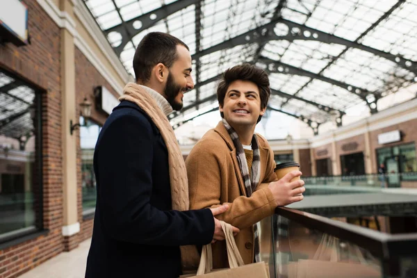 Sonriente gay hombre con café a ir mirando barbudo novio con compras bolsas cerca borrosa edificio con tiendas - foto de stock
