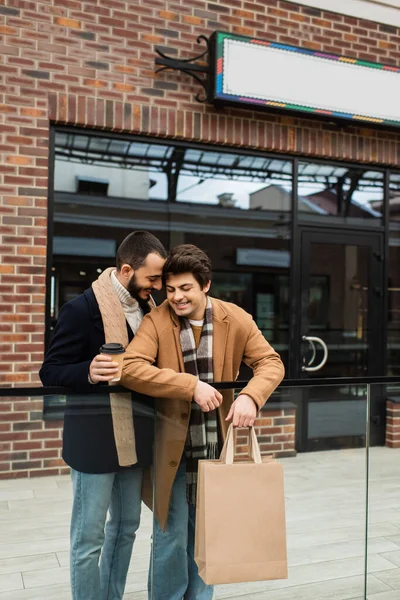 Moda gay parceiros com compras sacos e café para ir sorrindo perto de cerca de vidro e construção com vitrine — Fotografia de Stock