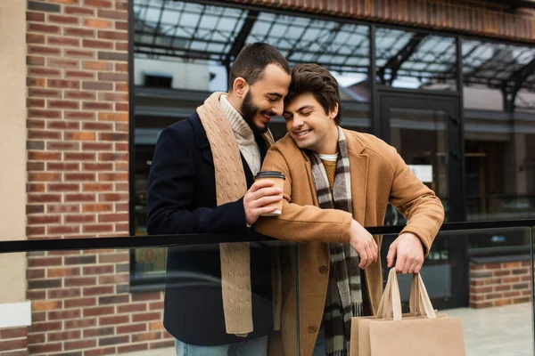 Heureux gay l'homme tenant à emporter boisson près de jeune copain avec shopping sacs et magasin sur fond — Photo de stock