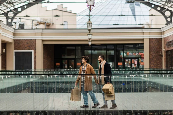 Full length of trendy homosexual couple with shopping bags waking near building with stores — Stock Photo