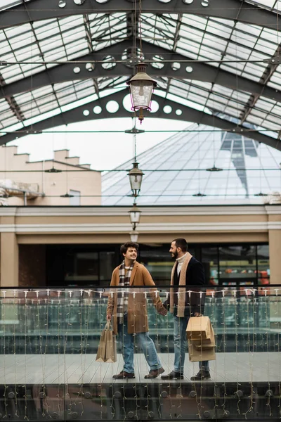 Stylish gay men with shopping bags holding hands and looking at each other under transparent roof outdoors — Stock Photo