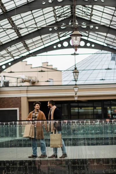 Full length of stylish gay partners with shopping bags walking under transparent roof near building with shops — Stock Photo
