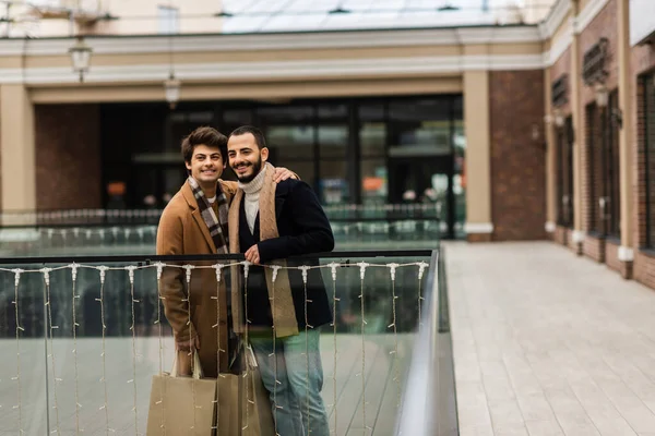 Trendy and happy gay men with shopping bags standing near glass fence and blurred building — Stock Photo