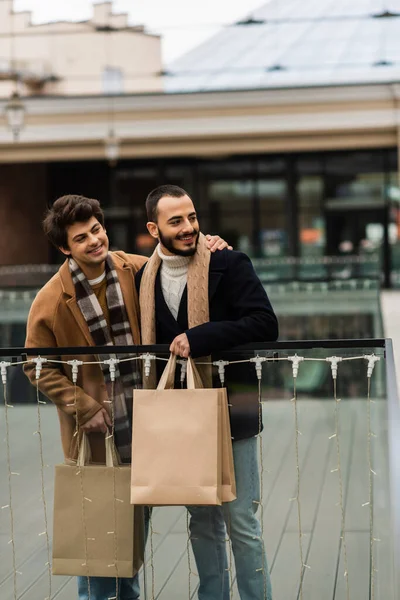 Pareja gay de moda con bolsas de compras sonriendo y mirando hacia otro lado cerca de la valla de vidrio al aire libre - foto de stock