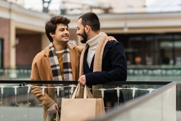 Счастливые геи в модных пальто и шарфах держат сумки с покупками и смотрят друг на друга на городской улице — стоковое фото