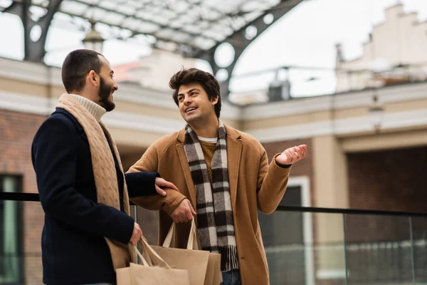 Giovane gay in plaid sciarpa punta con mano e parlando con barbuto fidanzato con shopping bags — Foto stock