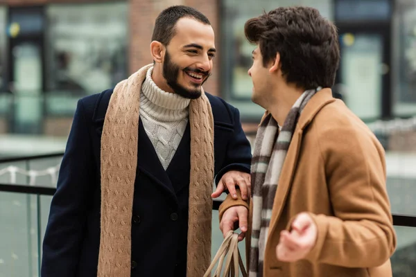 Bärtiger und fröhlicher schwuler Mann in schwarzem Mantel und Schal im Gespräch mit jungem Freund im Freien — Stockfoto