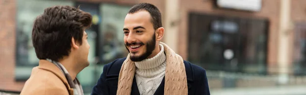 Barbuto uomo in sciarpa sorridente a giovane gay partner su blurred urbano strada, striscione — Foto stock