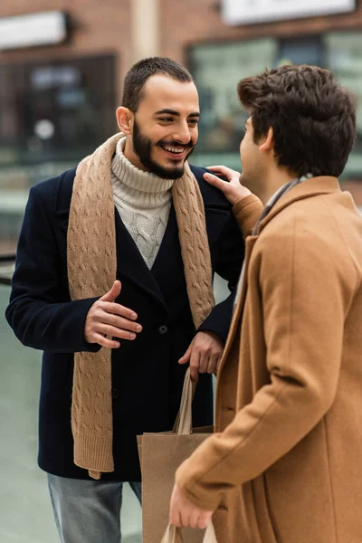 Fröhlicher bärtiger Schwuler in schwarzem Mantel, der Einkaufstüten hält, während er mit seinem Freund spricht, der seine Schulter berührt — Stockfoto
