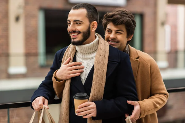 Бородатий гей у пальто та шарфі стоїть з виносним напоєм біля хлопця, обіймаючи його на відкритому повітрі — стокове фото