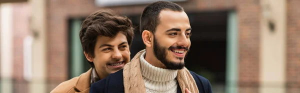 Joven y feliz gay hombre sonriendo cerca barbudo novio al aire libre, bandera — Stock Photo