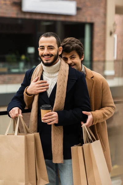 Barbu gay l'homme dans noir manteau tenant shopping sacs et papier tasse près de jeune copain étreinte lui à l'extérieur — Photo de stock