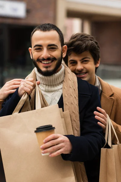 Весёлый гей обнимает бородатого парня с бумажной чашкой и пакетами для покупок — стоковое фото