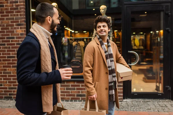 Улыбающийся гей с покупками смотрит на парня с бумажной чашкой возле магазина на городской улице — стоковое фото