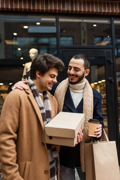Happy gay man holding shoebox near bearded boyfriend with paper cup and shopping bag at blurred showcase — Stock Photo