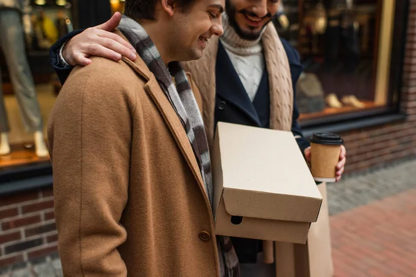 Smiling gay man with takeaway drink hugging boyfriend with shoebox near showcase on blurred background — Stock Photo