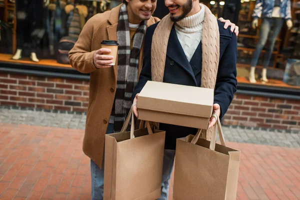 Corte vista de sorrindo gay homem com café para ir abraçando namorado com shoebox e compras sacos no o rua — Fotografia de Stock